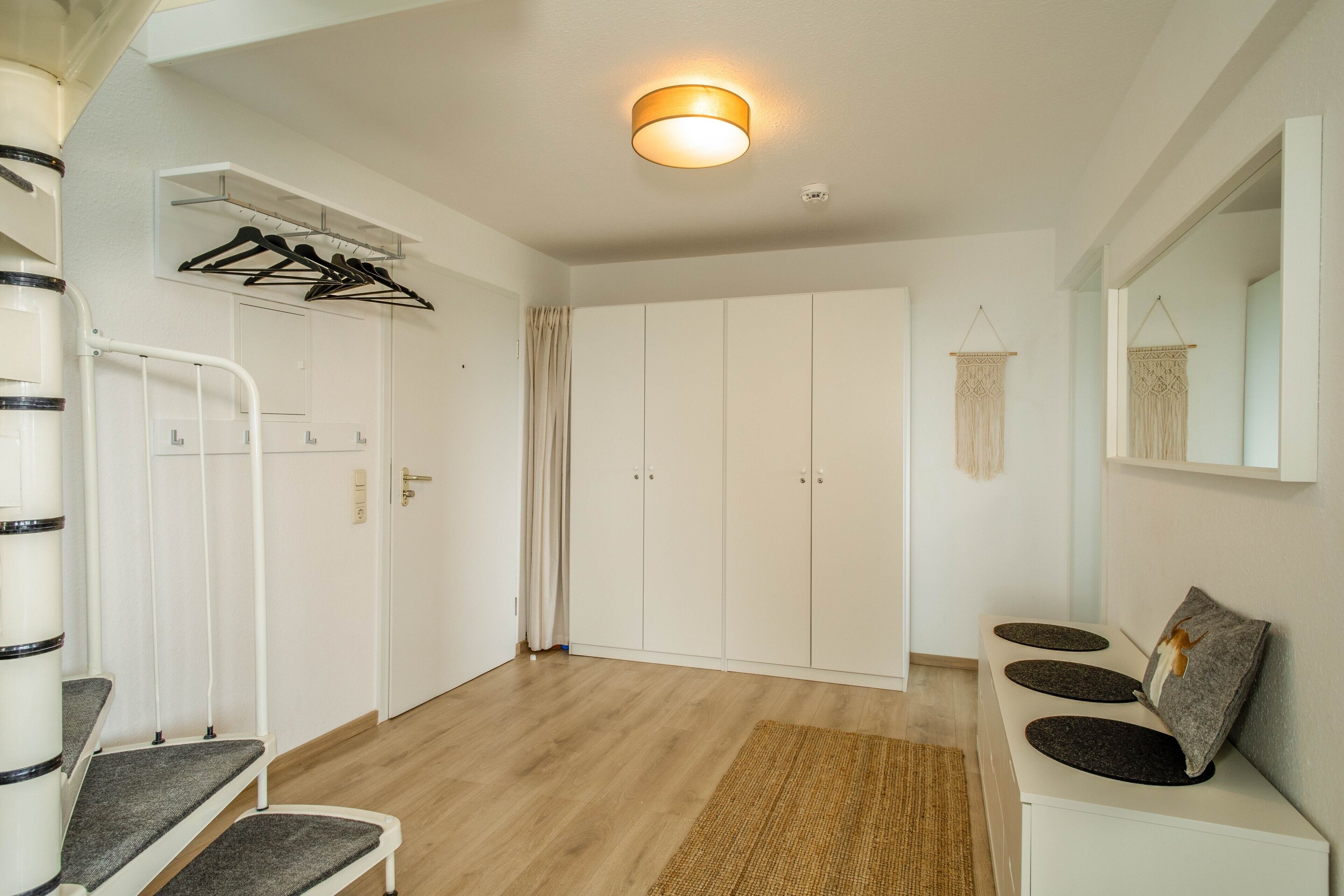 Ein Zimmer mit einem weißen Kleiderschrank, einer Bank, einem kleinen Tisch, einem Spiegel und einer Pendelleuchte.