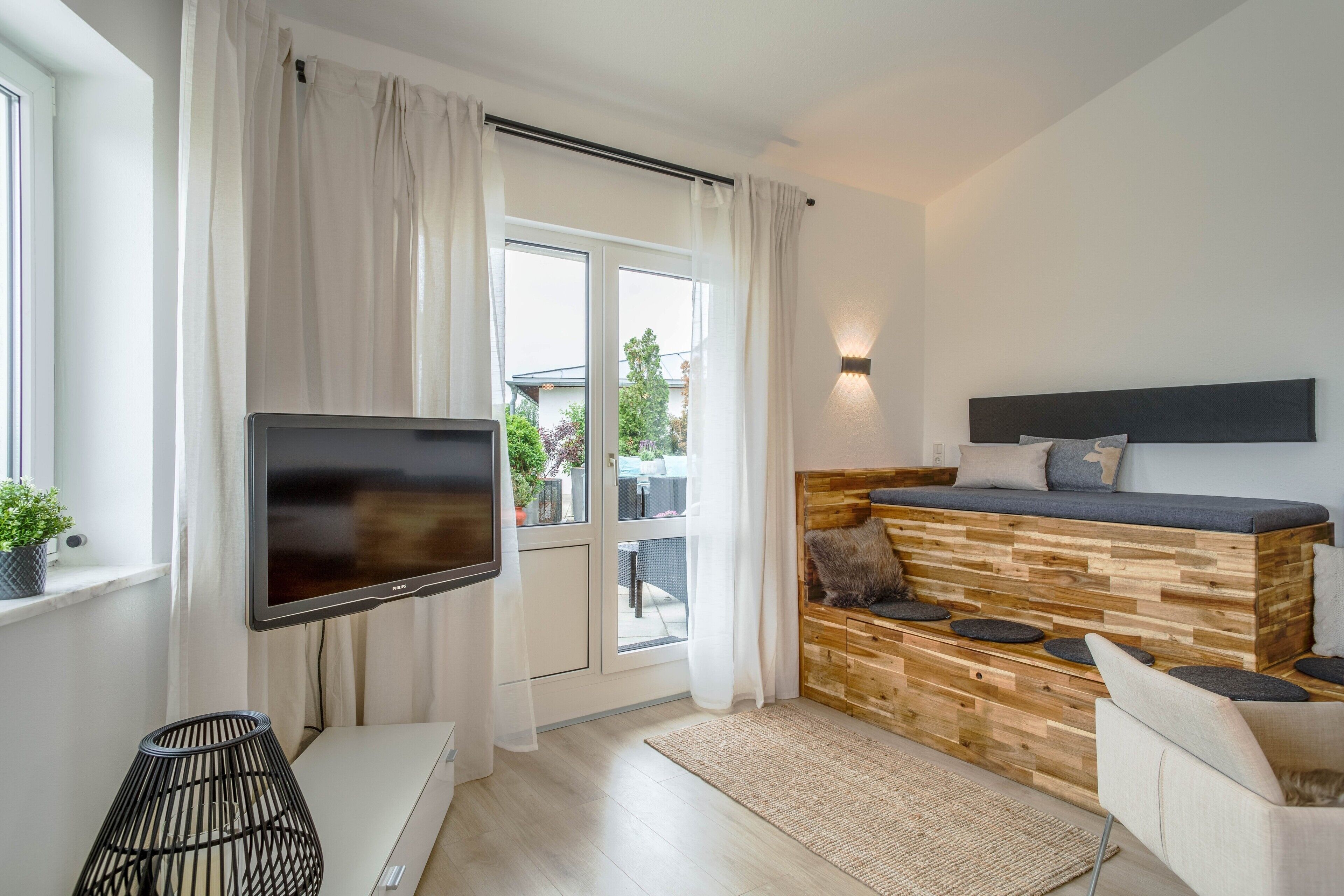 A modern living room with a flat-screen TV, a wooden bench with cushions, a sofa, and a large window with curtains.