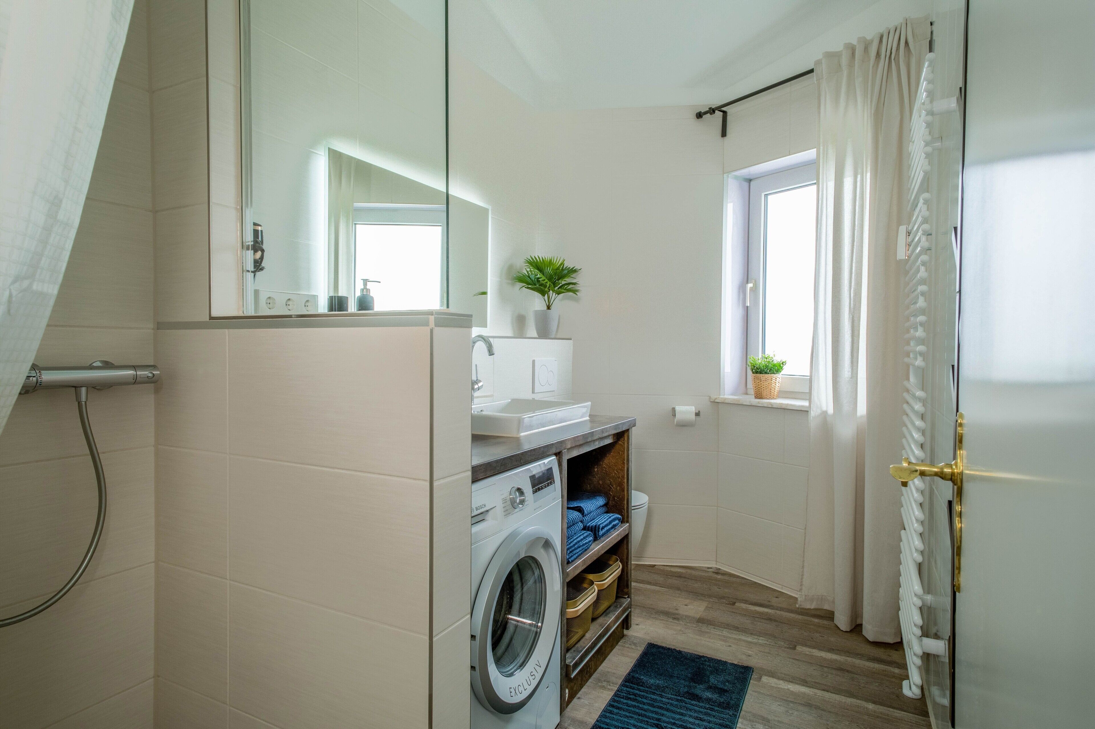 Ein kleines Badezimmer mit Waschmaschine, Waschbecken, Spiegel, Fenster mit Vorhängen und Heizkörper.