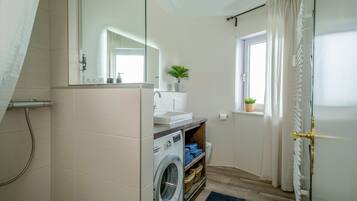 Ein kleines Badezimmer mit Waschmaschine, Waschbecken, Spiegel, Fenster mit Vorhängen und Heizkörper.