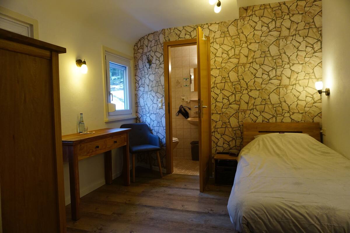 A bedroom with a wooden desk, a chair, a bed, and a bathroom visible through an open door.