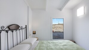 Een slaapkamer met een bed, nachtkastje, lamp en uitzicht op een rotsachtig landschap door een deur.
