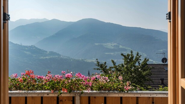 Uitzicht door een open deur met houten frame, met roze bloemen op de voorgrond en een bergachtig landschap op de achtergrond.