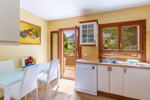 Une cuisine équipée d’un lave-vaisselle, une table à manger avec des chaises, un panier de fruits et un tableau accroché au mur.