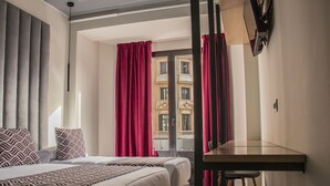 A hotel room with a bed, a desk, and a view of a building through a window with red curtains. - Hostal Juliette & Coworking (Madrid)