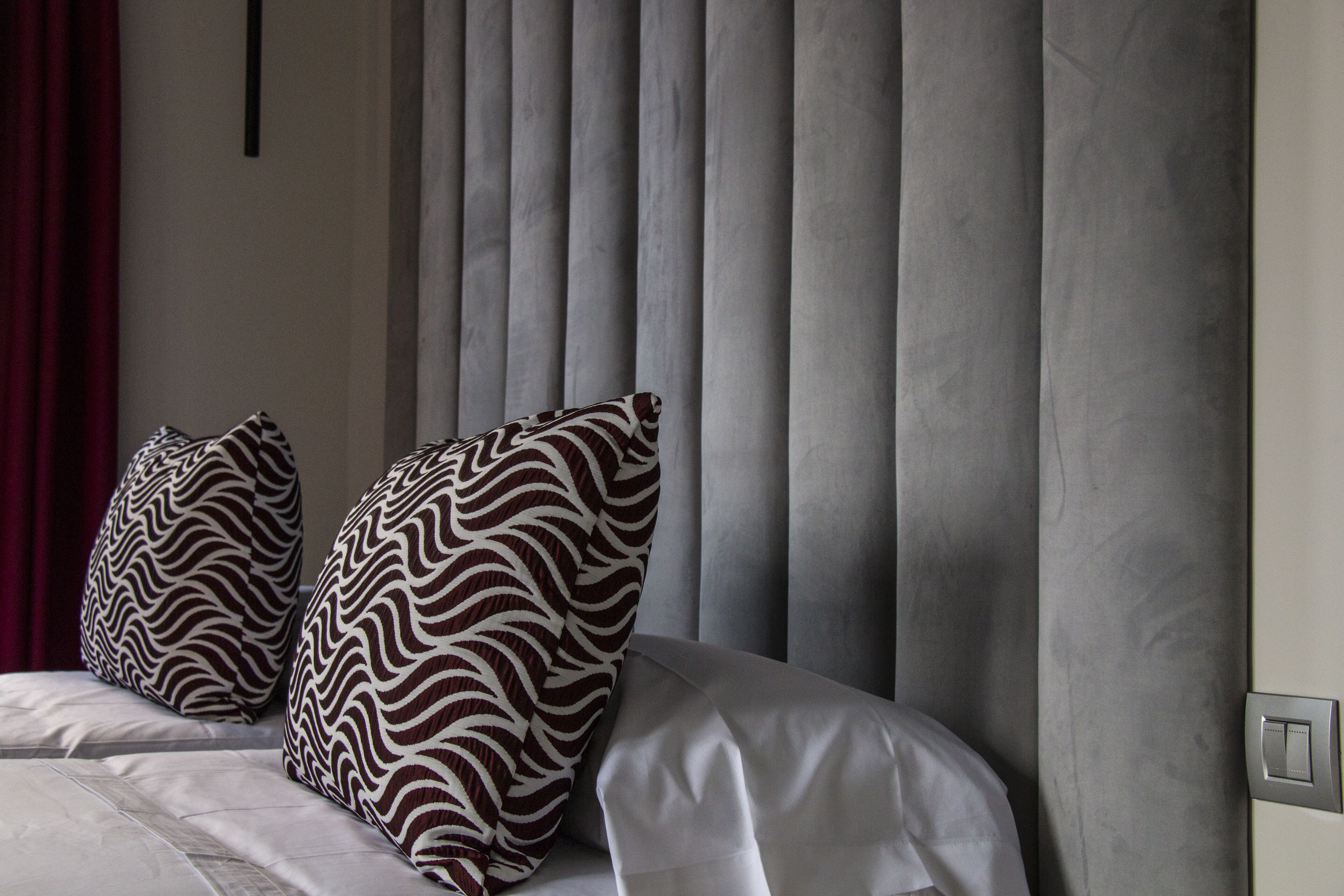 A bed with white bedding and two patterned pillows, a grey curtain, and a light switch on the wall.