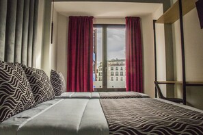 A hotel room with a large bed, two pillows, a window with red curtains, and a view of buildings outside. - Hostal Juliette & Coworking (Madrid)
