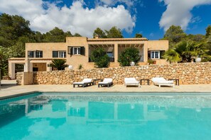 A poolside view of a Mediterranean-style villa with a stone wall, multiple lounge chairs, and a clear blue pool.
