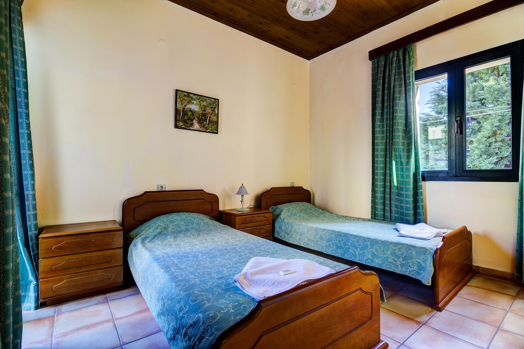 A room with two wooden beds, a nightstand with a lamp, a small painting on the wall, and a window with curtains.