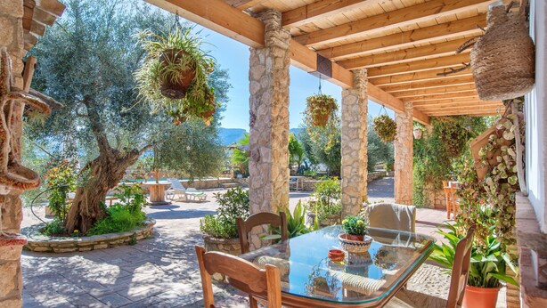 Une terrasse extérieure couverte, dotée d’une table en verre, de piliers en pierre et de plantes suspendues.
