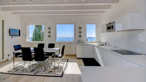 Una cocina moderna con muebles blancos, un comedor con mesa y sillas, y ventanas amplias que dan a vista al mar.