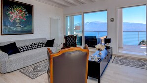 Una sala de estar con sofá, sillón y una mesa con adornos. Hay un cuadro en la pared y vistas al mar a través de ventanas amplias.