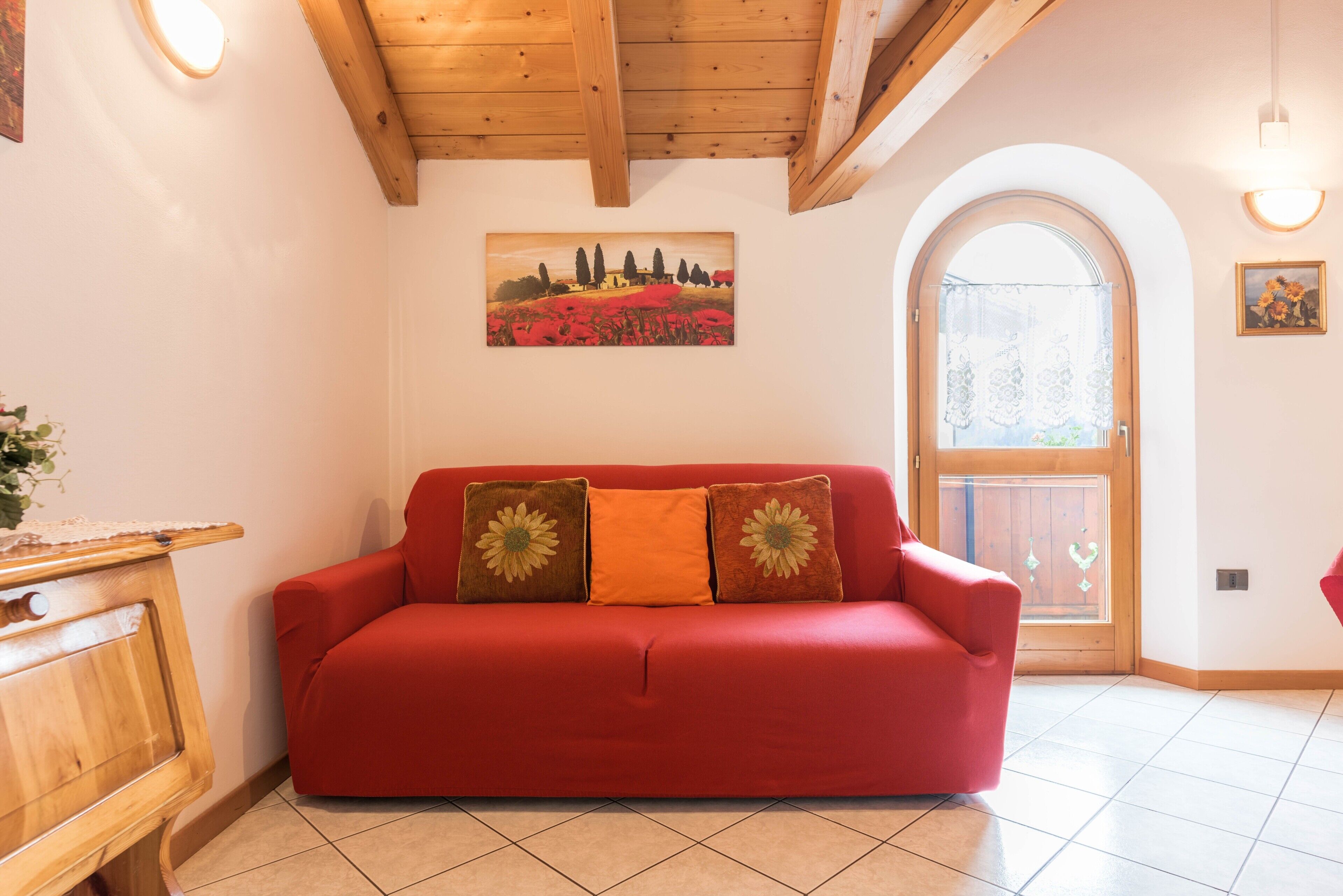 Un sofá rojo con cojines estampados, un mueble de madera, un cuadro de un paisaje y una puerta de madera con un ventanal.