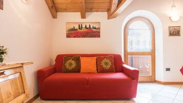 Un sofá rojo con cojines estampados, un mueble de madera, un cuadro de un paisaje y una puerta de madera con un ventanal.
