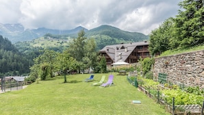 A mountain lodge with a stone wall, a grassy lawn, and a view of the surrounding mountains. - Holiday Apartment "Bamby - Vioz" with Balcony, Garden & Mountain View (Cogolo di Pejo)