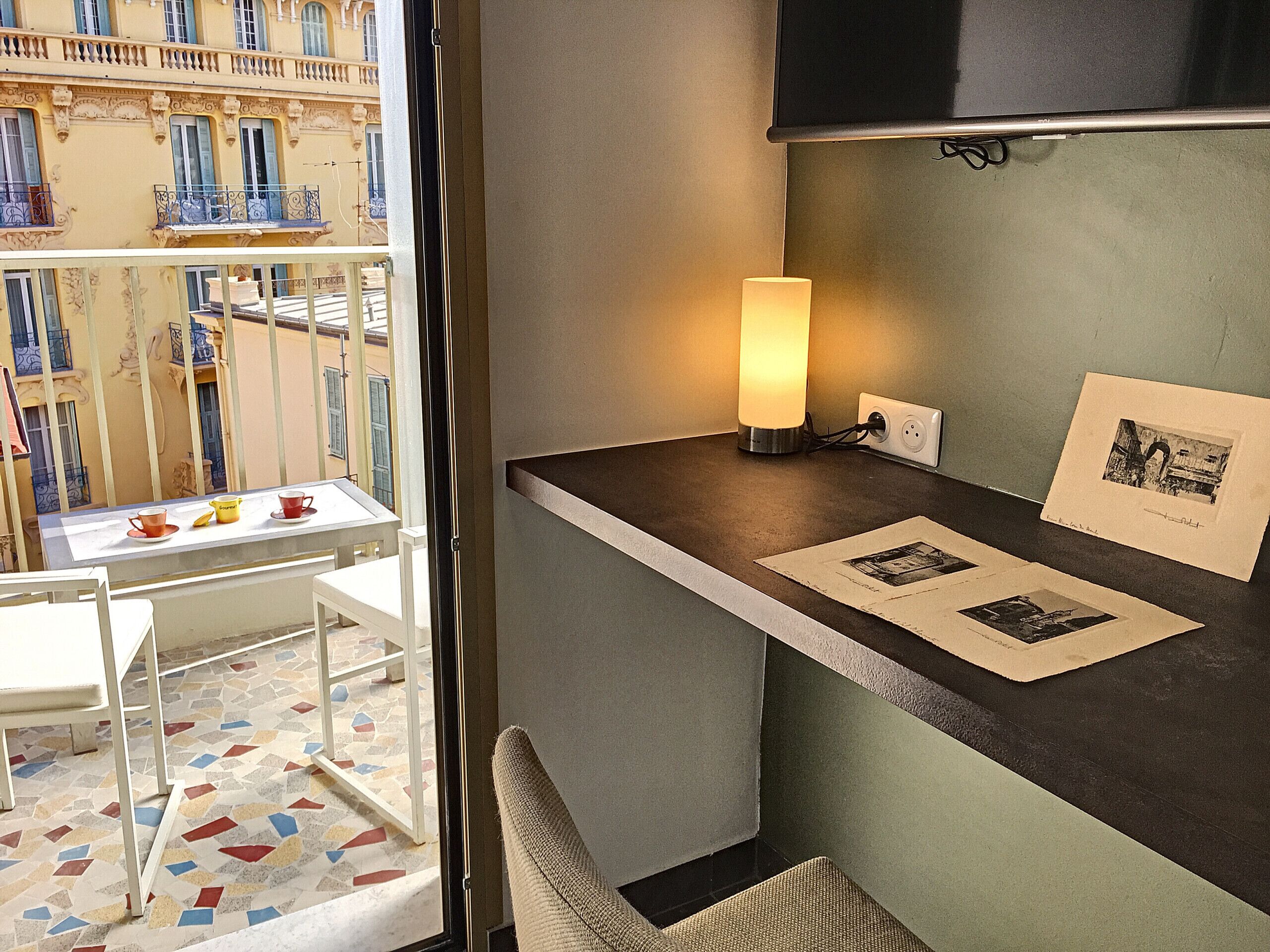 A balcony with a table and chairs, a standing lamp, and a wall-mounted shelf with framed pictures.