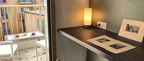 A balcony with a table and chairs, a standing lamp, and a wall-mounted shelf with framed pictures.
