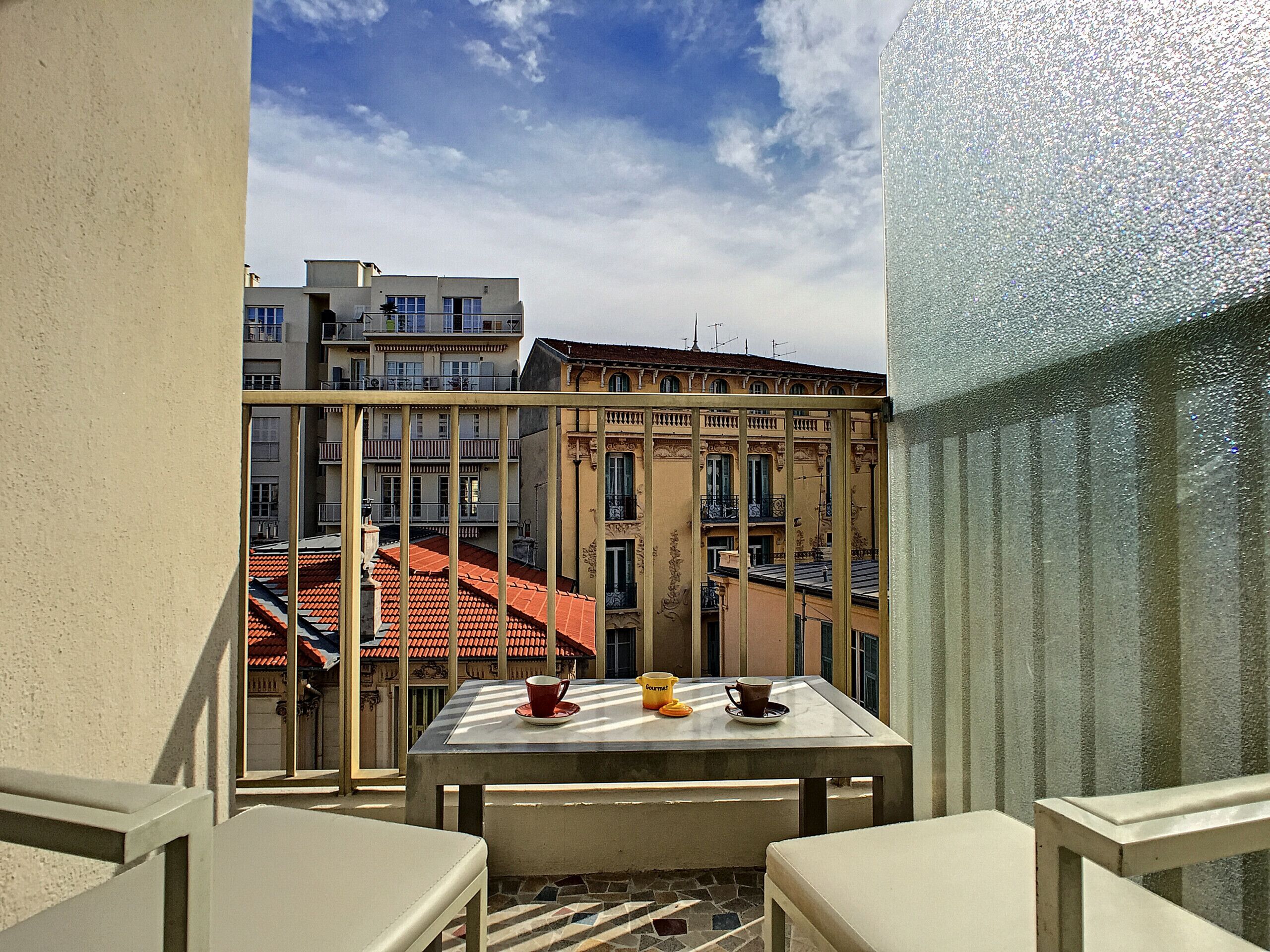 Un balcon avec une table et des chaises, donnant sur un paysage urbain avec des bâtiments et un ciel dégagé.