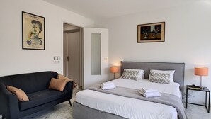 Habitación de hotel con cama, sofá, mesita de noche, lámpara y dos cuadros en las paredes.