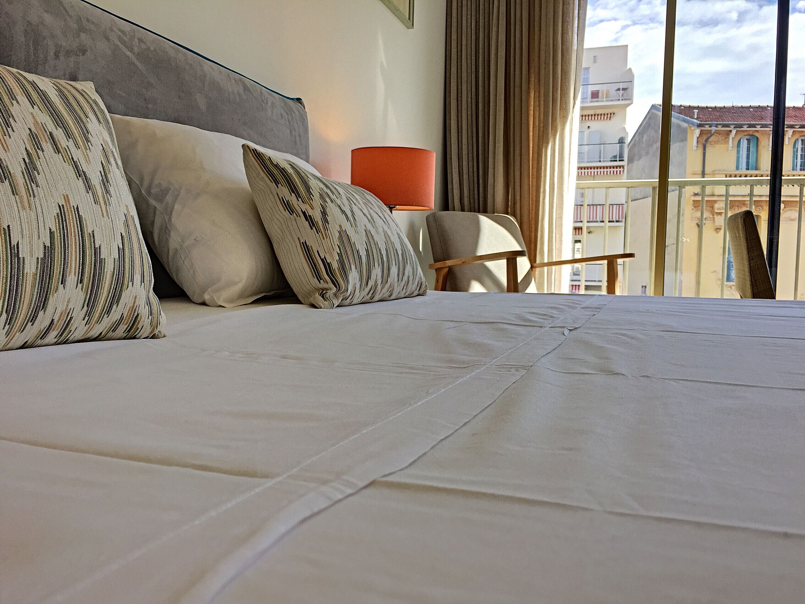 Un lit bien fait, agrémenté d’oreillers à motifs, une chaise en bois et une vue sur les immeubles par la fenêtre.