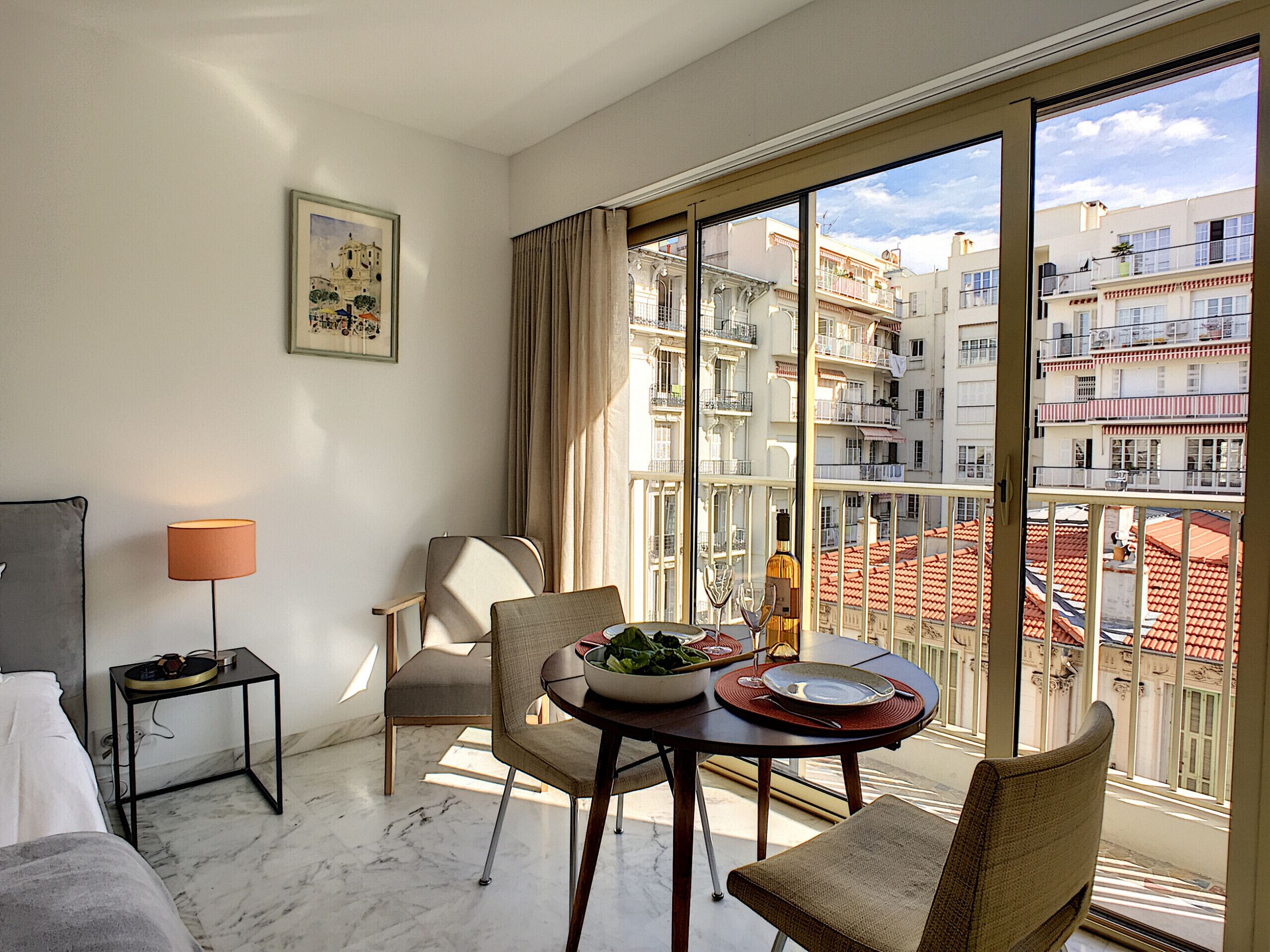 A modern hotel room with a dining area, large windows, and a view of the cityscape.