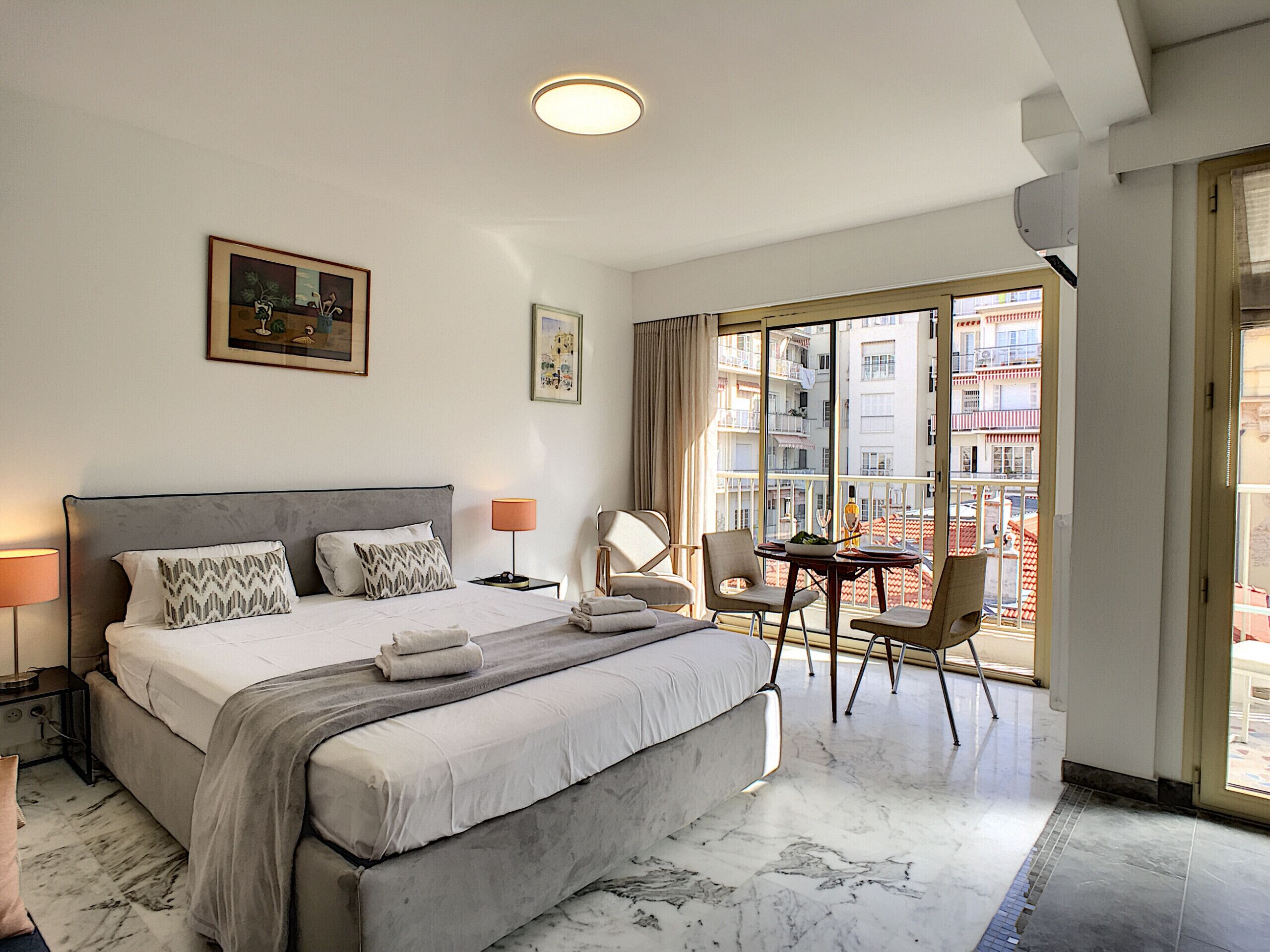 Habitación de hotel con una cama grande, una pequeña mesa con sillas, un balcón con vistas a edificios y una lámpara en el techo.