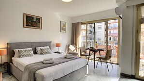 A hotel room with a large bed, a small table with chairs, a balcony with a view of buildings, and a ceiling light. - N&J - BERLIOZ SUITE - Central - Close sea (Niza)