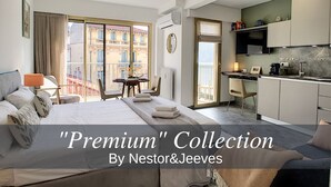 A hotel room with a bed, a desk, a chair, a small table, and a view of buildings through large windows. - N&J - BERLIOZ SUITE - Central - Close sea (Niza)