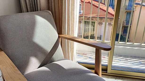 Una habitación con un sillón gris, una mesita de madera y vistas a un edificio con balcones y un tejado de tejas rojas a través de una ventana con barandilla de metal.