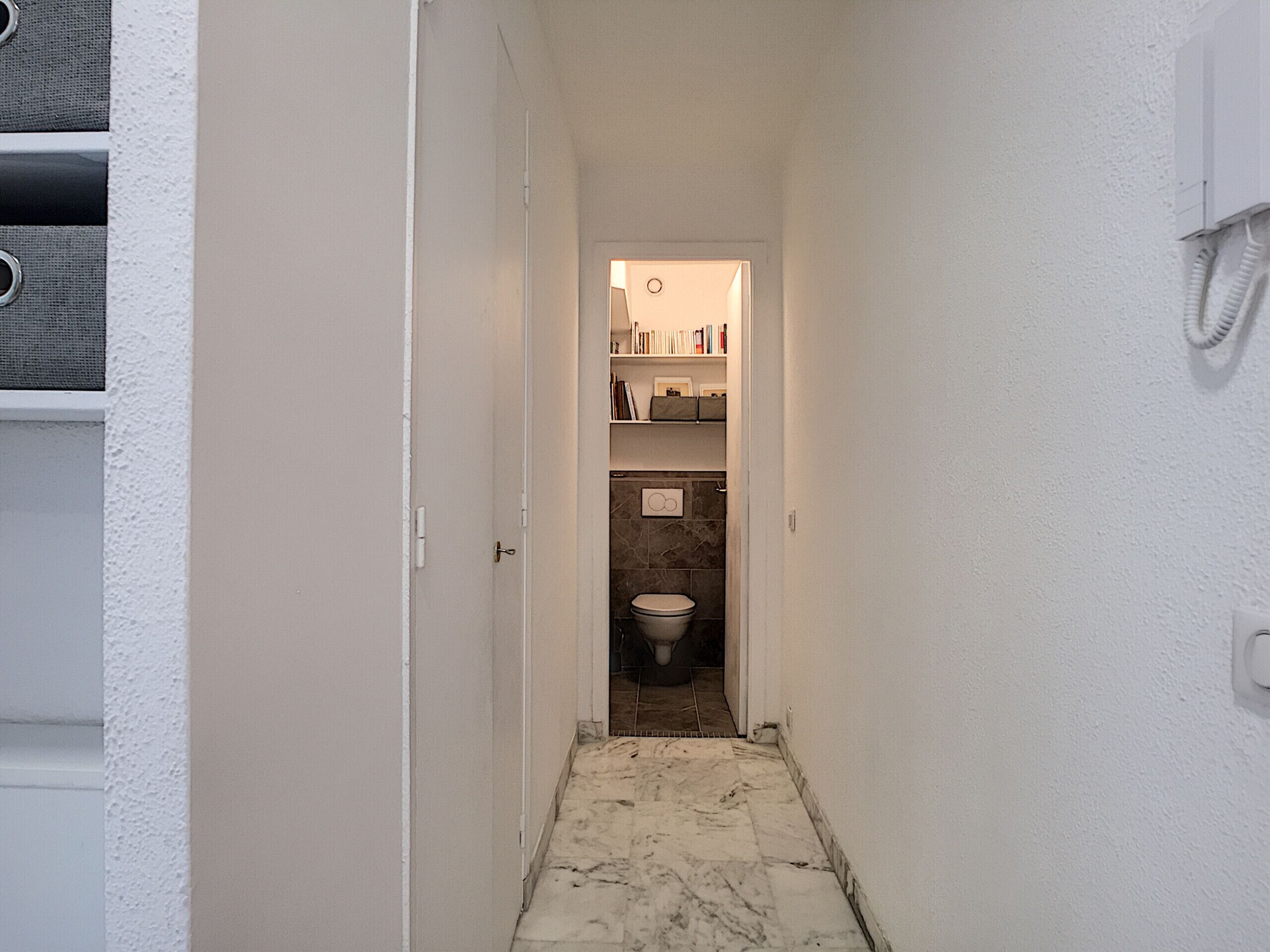 A narrow hallway leading to a bathroom with a toilet, shelves with books, and a white wall with a coiled cord.
