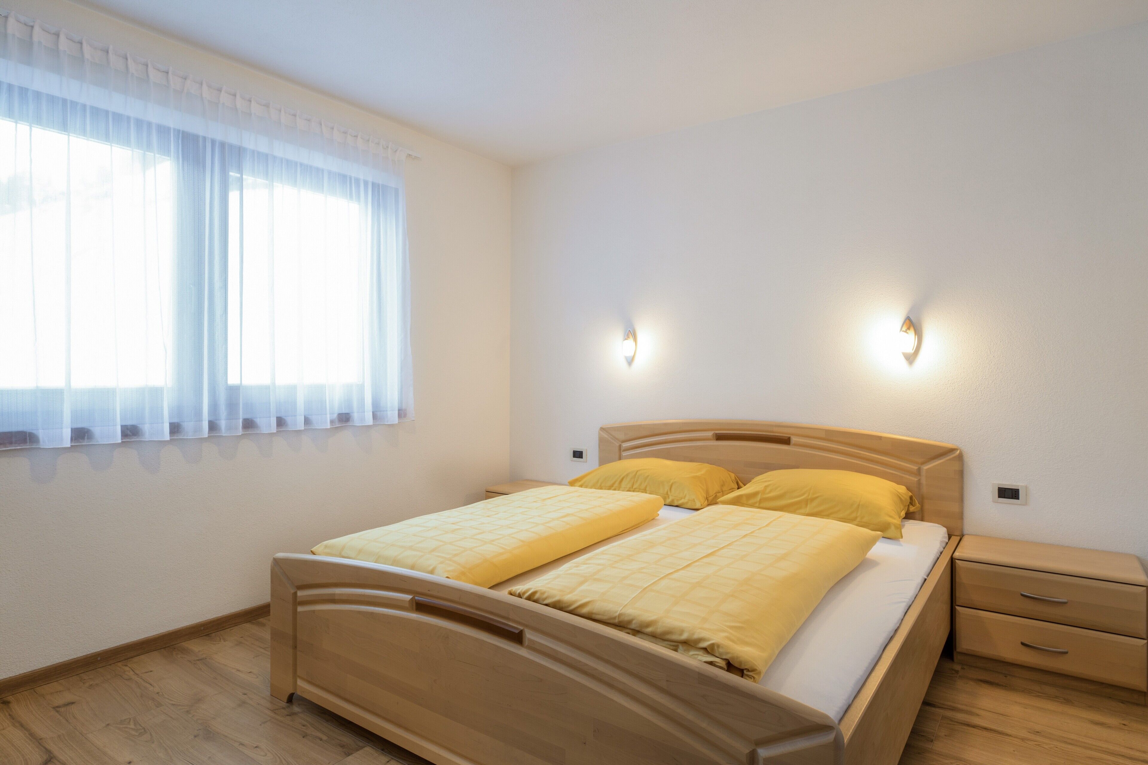 A bedroom with a wooden bed, two bedside tables, a nightstand, and a window with sheer curtains.
