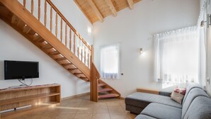 A living room with a wooden staircase, a flat-screen TV, a grey sofa, and a window with curtains. - Idyllic Apartment "Kronplatz" with Balcony, Garden and Wi-Fi (San Martino in Badia)
