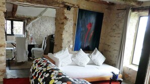 Une pièce rustique avec des poutres apparentes, un lit recouvert de linge de maison blanc, un canapé aux motifs colorés et un tableau accroché au mur.
