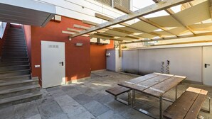 Un porche cubierto con una mesa de madera y bancos, una puerta y una escalera que lleva a un nivel superior.
