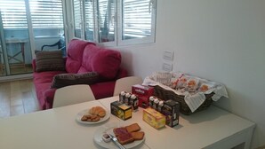 Una sala de estar con un sofá rojo, una mesa de comedor con cosas para el desayuno, y una ventana con persianas.
