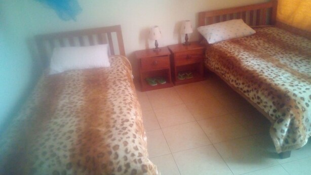 A room with two beds, each with a leopard print bedspread, wooden headboards, and matching pillows. There are bedside tables with lamps and a small shelf with slippers.