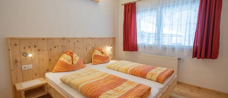 Ein Zimmer mit zwei Betten, einem Holz-Kopfteil, einem kleinen Nachttisch, einem Fenster mit roten Vorhängen und einer Heizung.