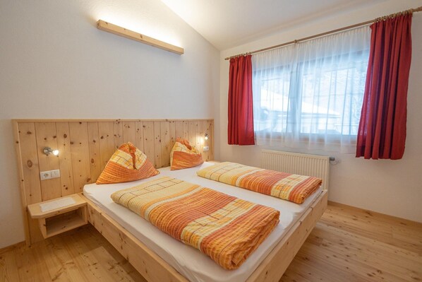 Une chambre avec deux lits, une tête de lit en bois, une petite table de chevet, une fenêtre avec des rideaux rouges et un radiateur.