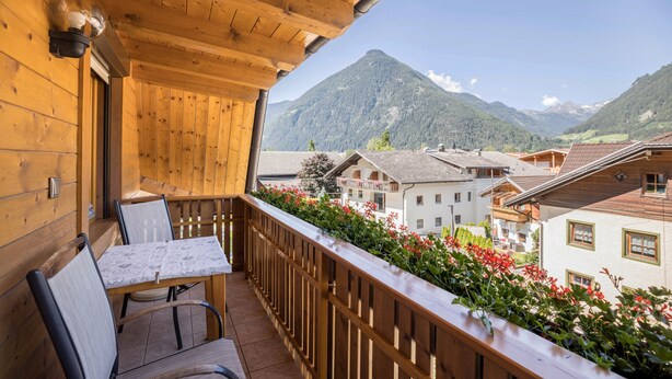 Ein Holzbalkon mit Tisch und Stühlen, der auf eine gebirgige Landschaft mit Gebäuden blickt.