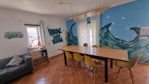 Een eetgedeelte met een houten tafel en gele stoelen, een grijze bank en een muurschildering met een golfmotief.