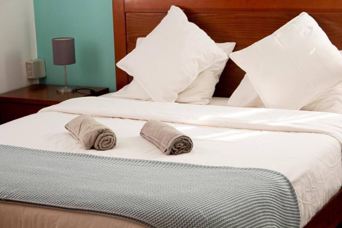 A neatly made bed with white linens and two rolled towels, a wooden headboard, and a bedside table with a lamp.