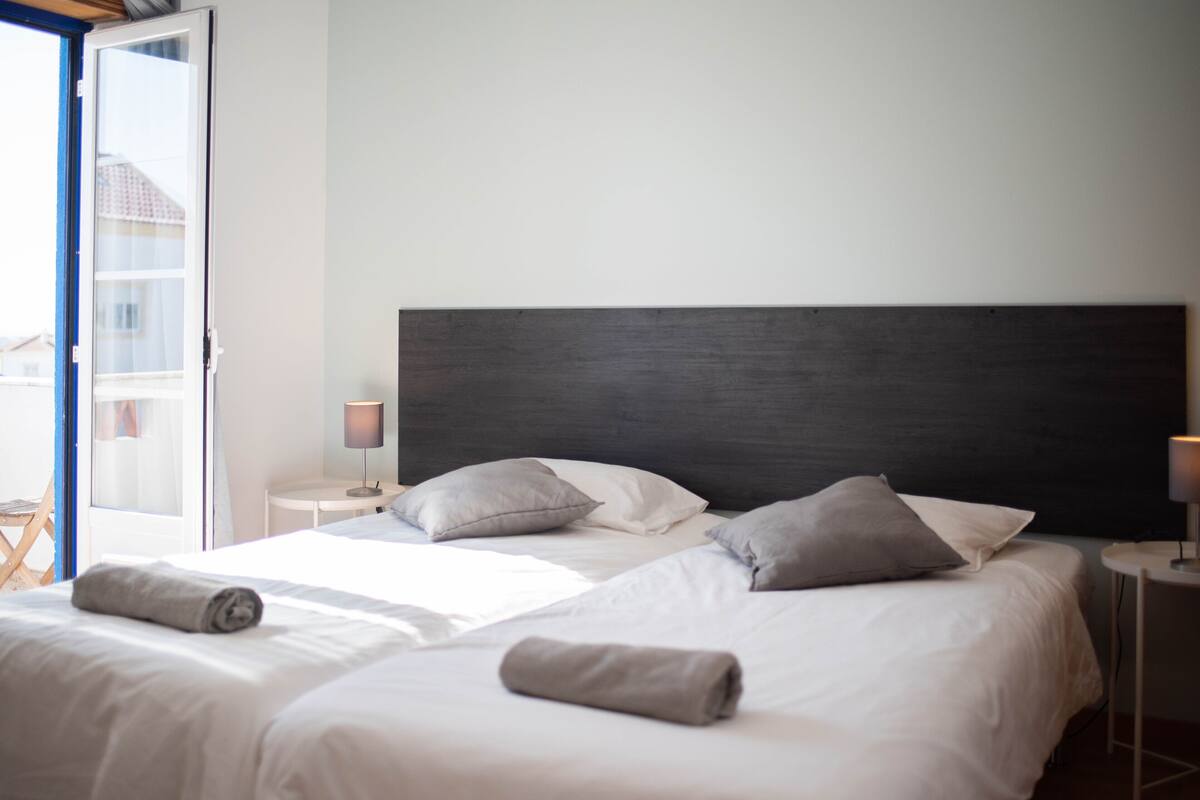A hotel room with two beds, a large dark headboard, and a view of the outside.