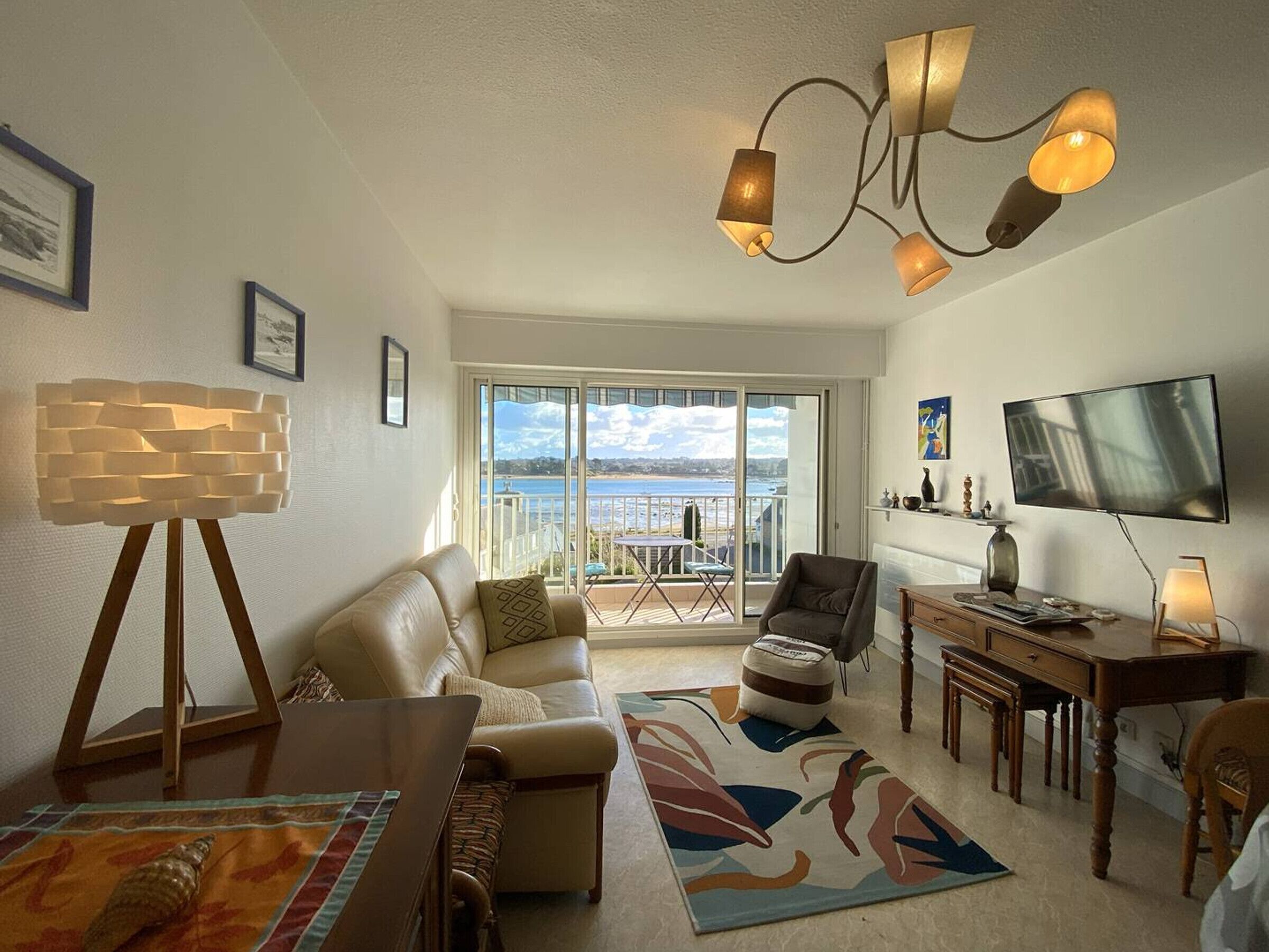 A living room with a sofa, a dining table, a TV, and a balcony with a view of the sea.