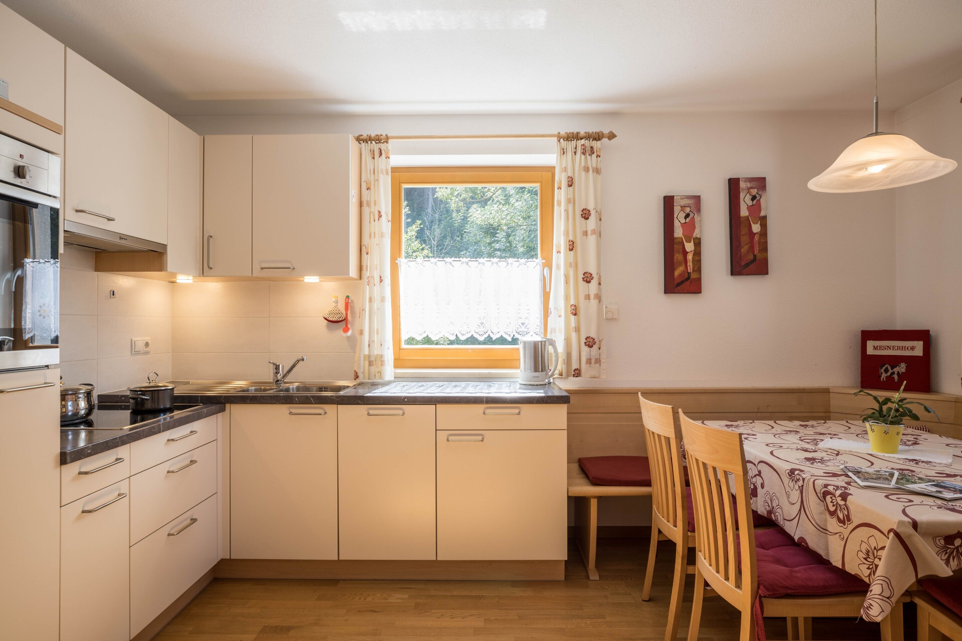 Eine kleine Küche mit Fenster, Holzschränken, einem Esstisch mit Stühlen und einer Hängeleuchte.