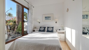 Ein Schlafzimmer mit einem großen Bett, zwei Nachttischen, einem Fenster mit Blick auf Pflanzen und einen Palmenbaum sowie einem gerahmten Bild an der Wand.