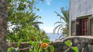 Eine Steinsäule mit tropischer Bepflanzung, ein Gebäude mit Fensterläden und Blick auf das Meer.