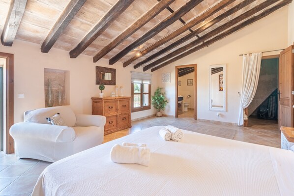 A spacious bedroom with a large bed, wooden beams, a wooden dresser, and a potted plant.