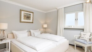 A hotel room with two beds, a bedside table with a lamp, a chair with a cushion, a framed picture on the wall, and a window with curtains. - Uhlenhof E from A&M - Uhlenhof E (Kampen (Sylt))