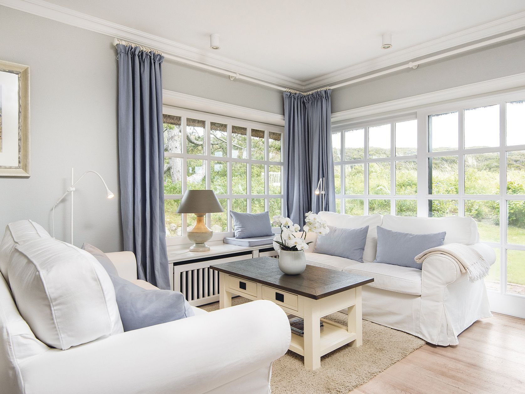 A spacious living room with a white sofa set, a wooden coffee table, and a view of greenery through large windows.