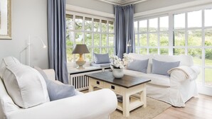 A spacious living room with a white sofa set, a wooden coffee table, and a view of greenery through large windows. - Uhlenhof E from A&M - Uhlenhof E (Kampen (Sylt))